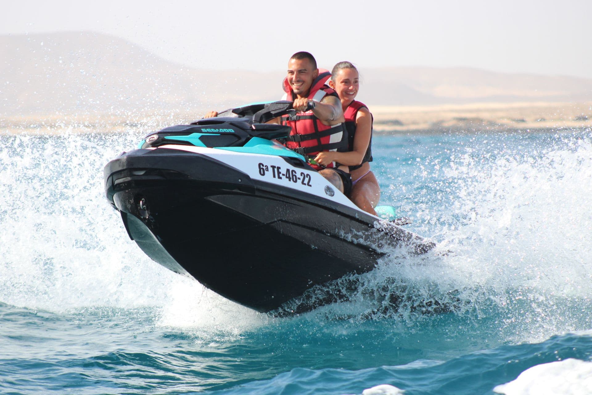 Laughing couple on a turquoise Sea-Doo with the pale sands of the Corralejo dunes behind