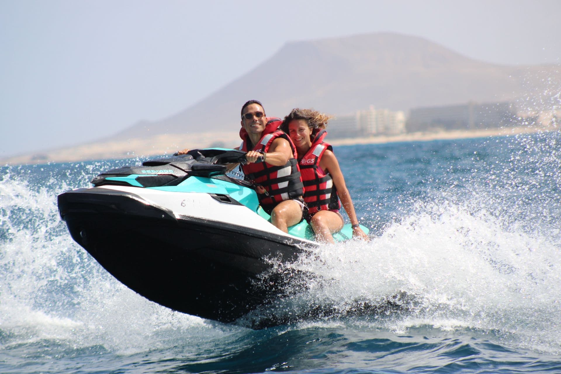 Smiling couple on a turquoise Sea-Doo with the volcanic mountain above the Corralejo dunes coastline behind them