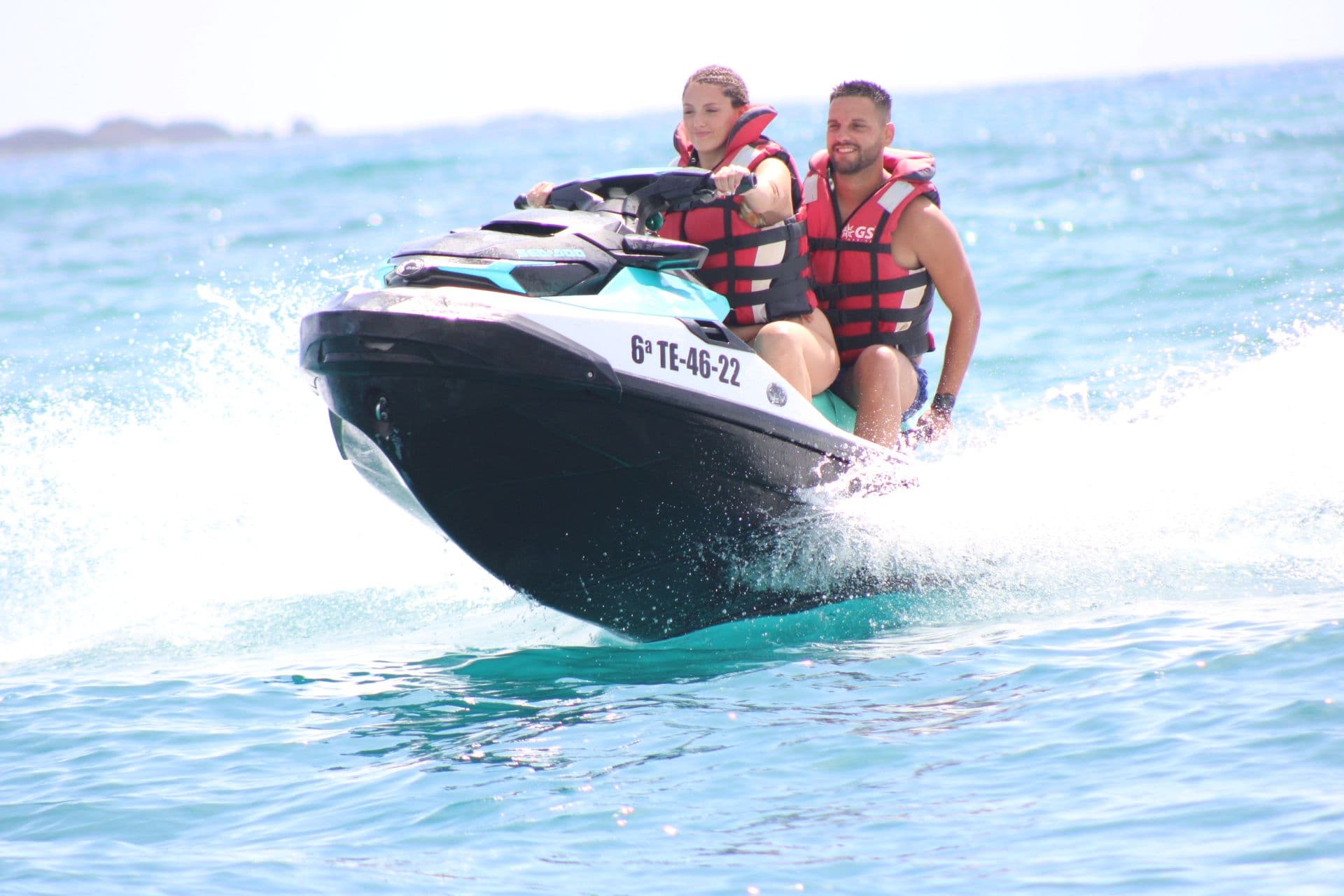 Smiling couple riding a turquoise and white jet ski straight towards the camera through turquoise water