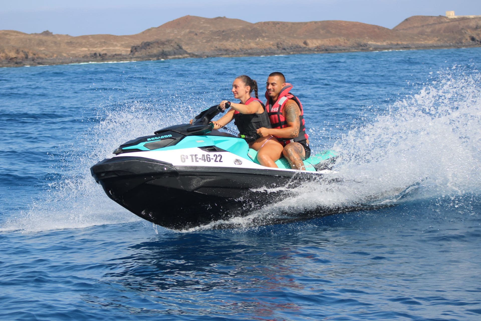 Couple on a turquoise jet ski with the low volcanic coastline of Isla de Lobos behind them