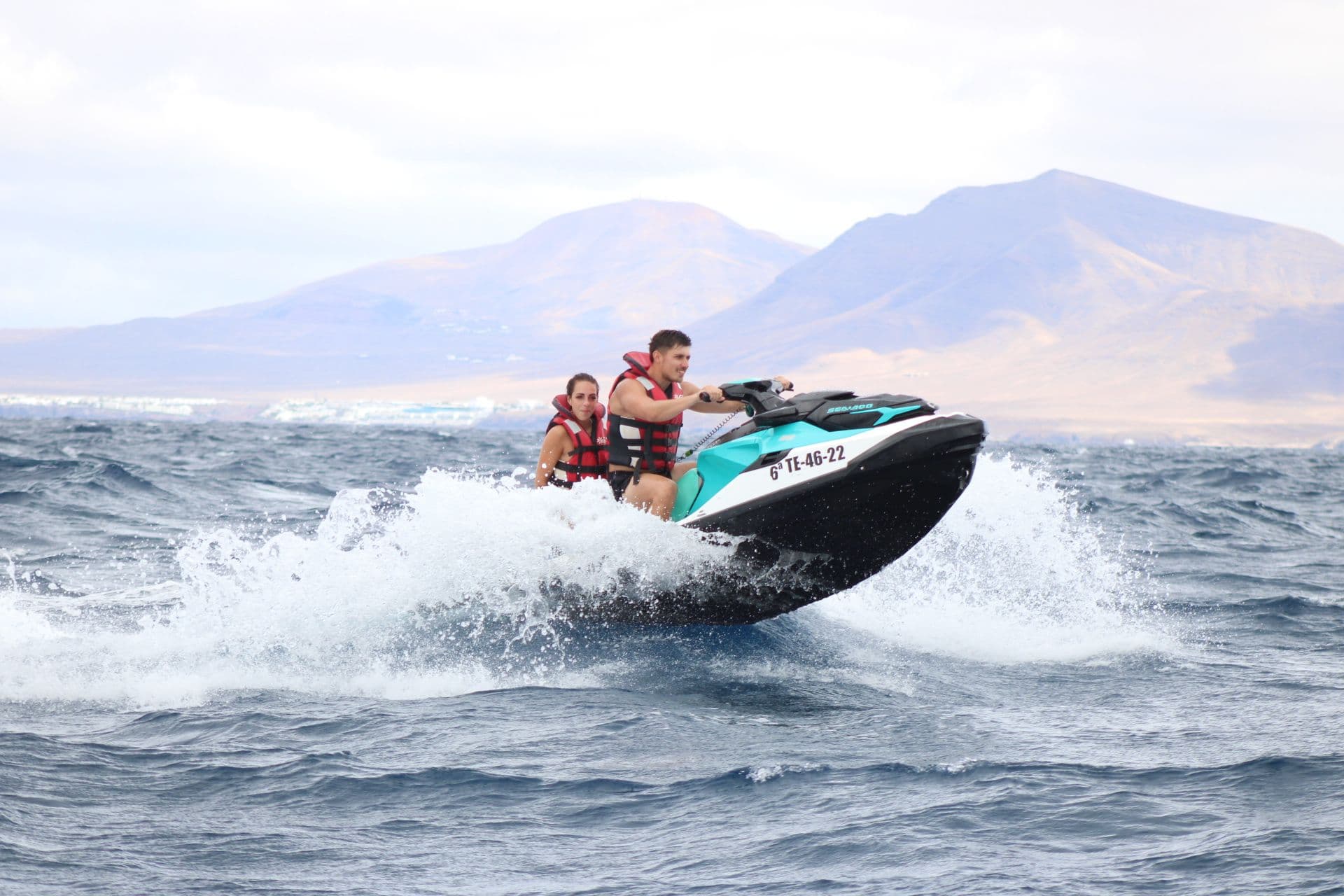 Couple riding a turquoise jet ski across the strait with the mountains of Lanzarote on the horizon