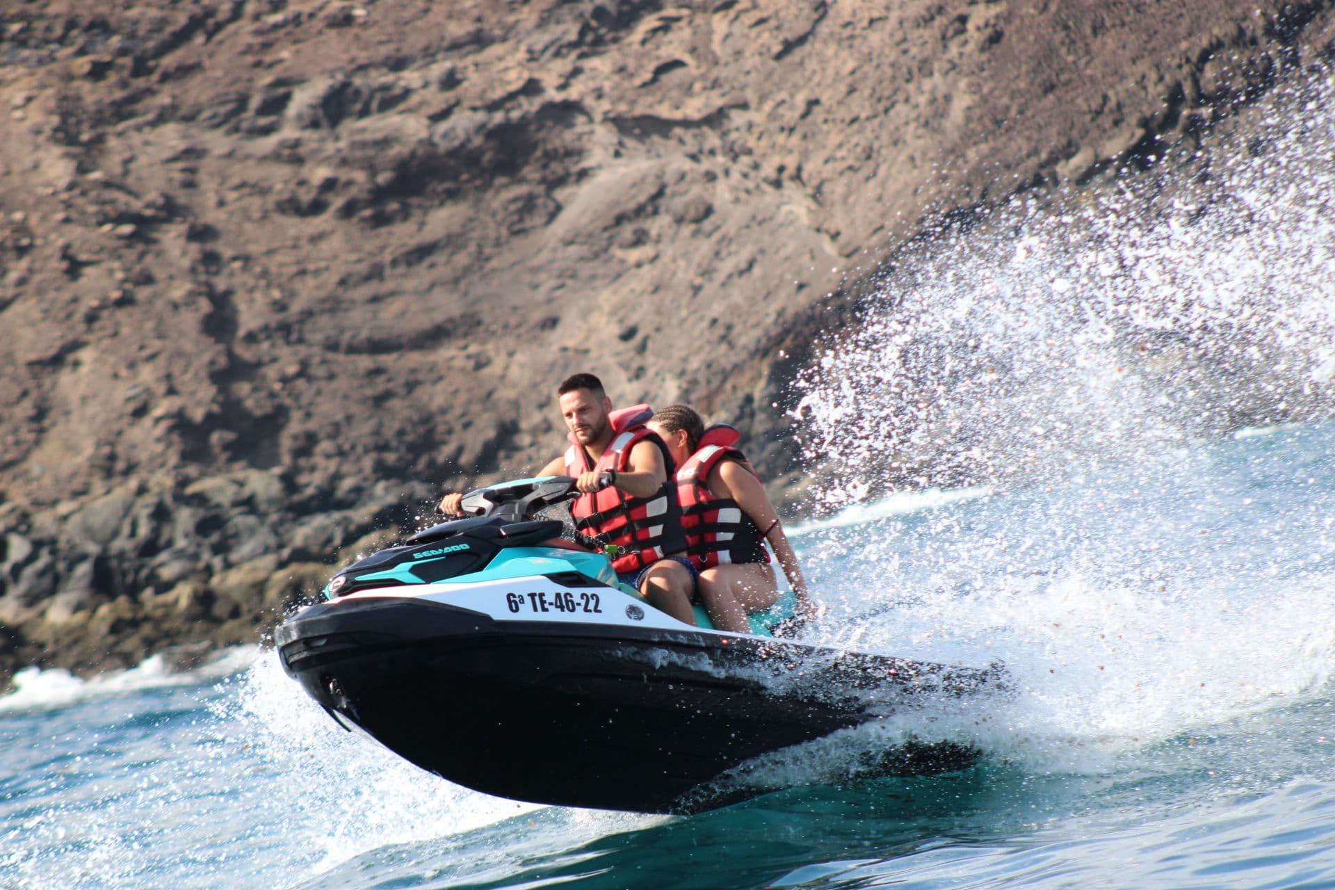 Couple riding past the volcanic lava rock coast of Isla de Lobos, with white spray off the jet ski