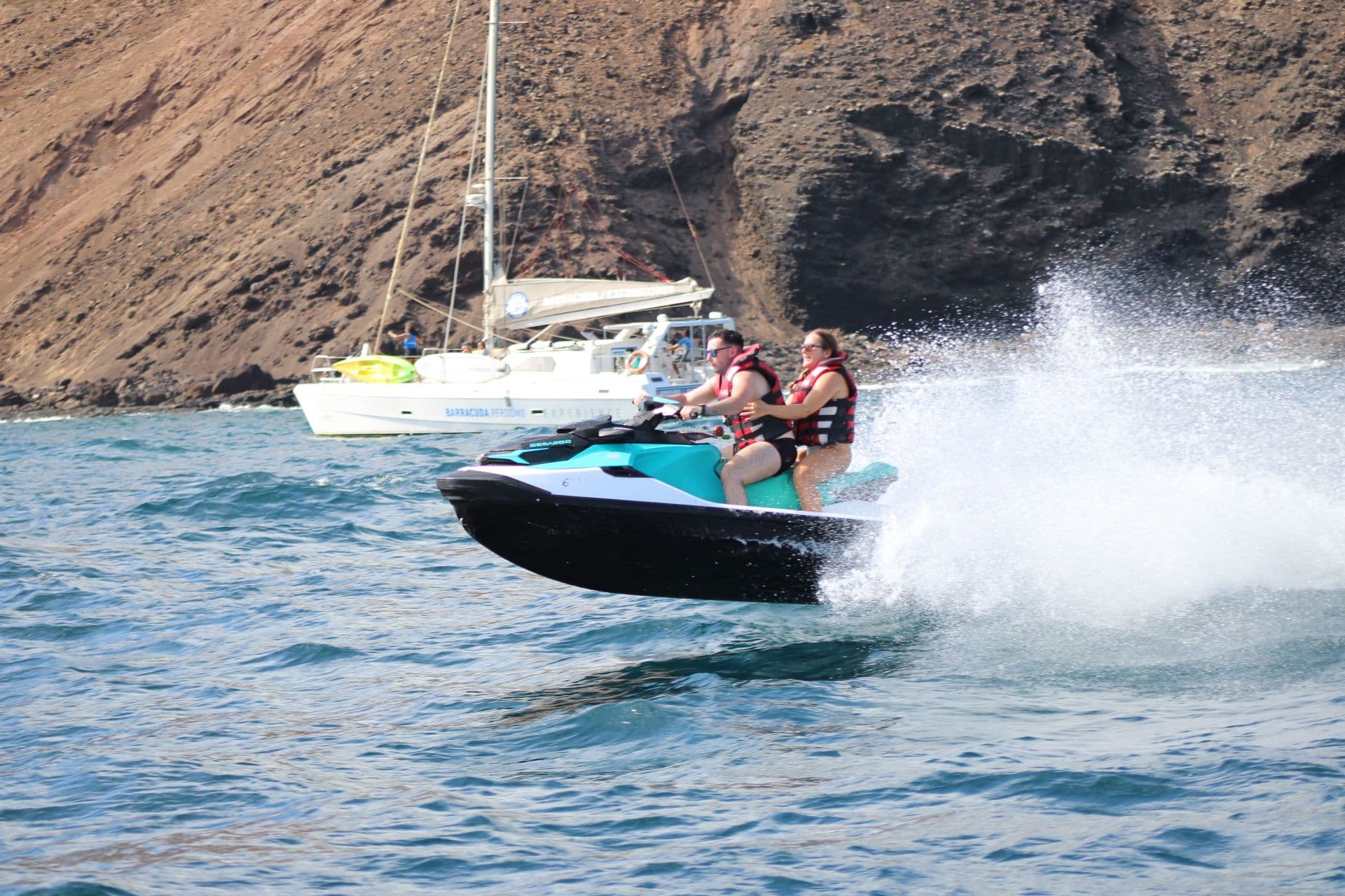 Couple on a turquoise jet ski below the red volcanic cliffs of Isla de Lobos, with a sailboat anchored nearby