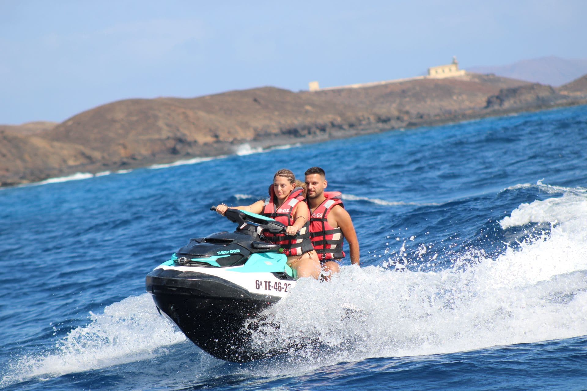 Smiling couple cutting through waves on a turquoise jet ski off Isla de Lobos, with the Punta Martiño lighthouse on the hill behind