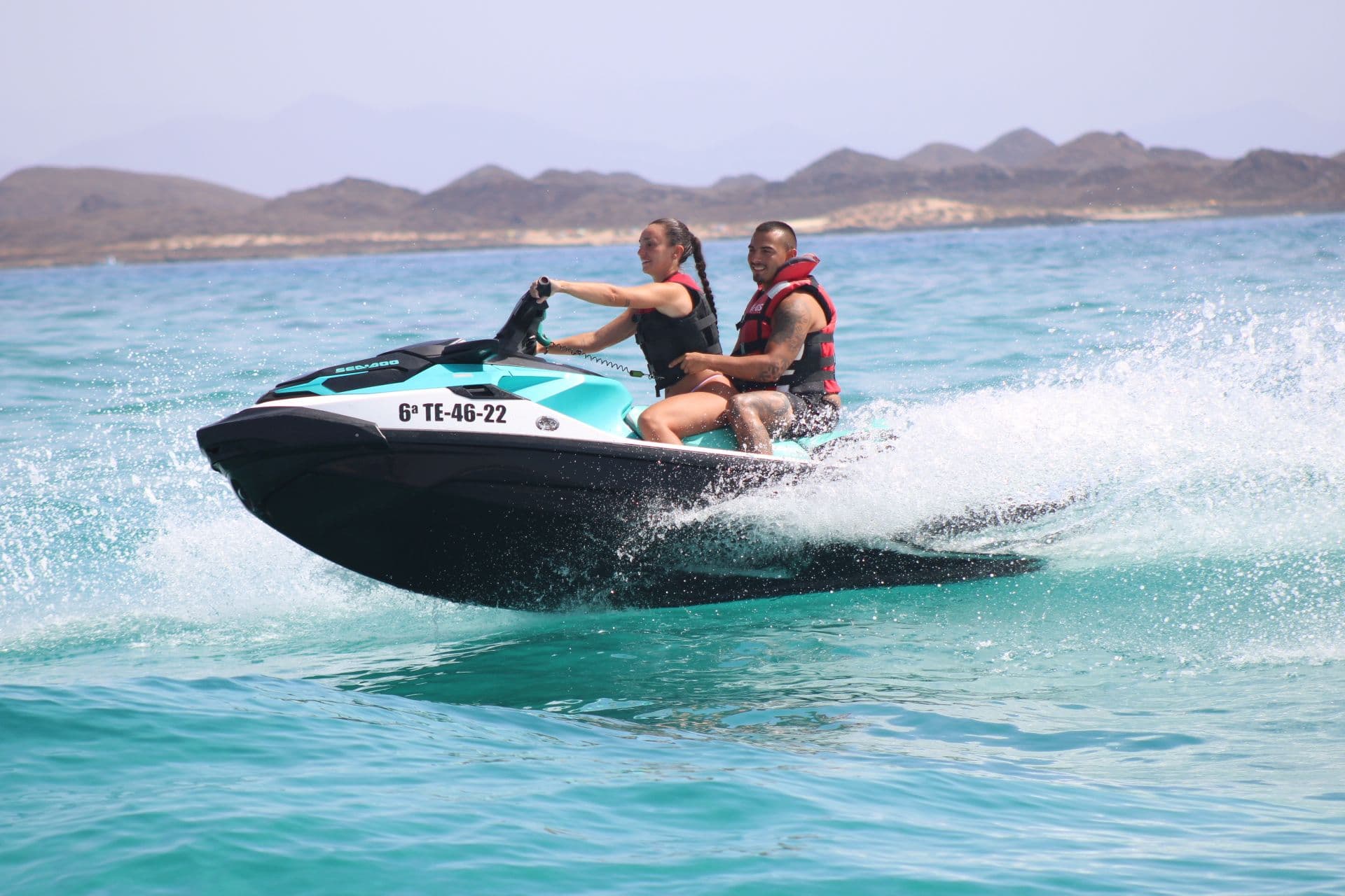 Couple on a turquoise jet ski over clear turquoise water in front of the hills of Isla de Lobos