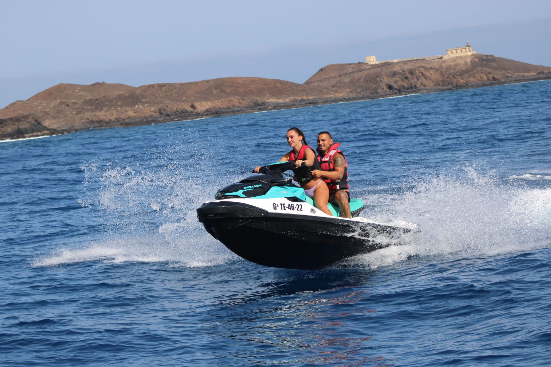 Couple riding a turquoise Sea-Doo jet ski with the Punta Martiño lighthouse on the Isla de Lobos headland behind them