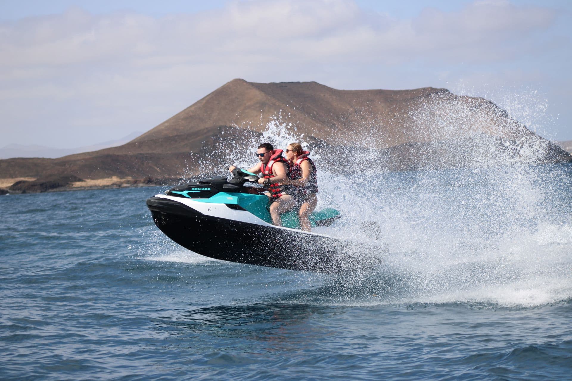 Couple on a jet ski in a burst of white spray with the volcanic cone of Isla de Lobos in the background