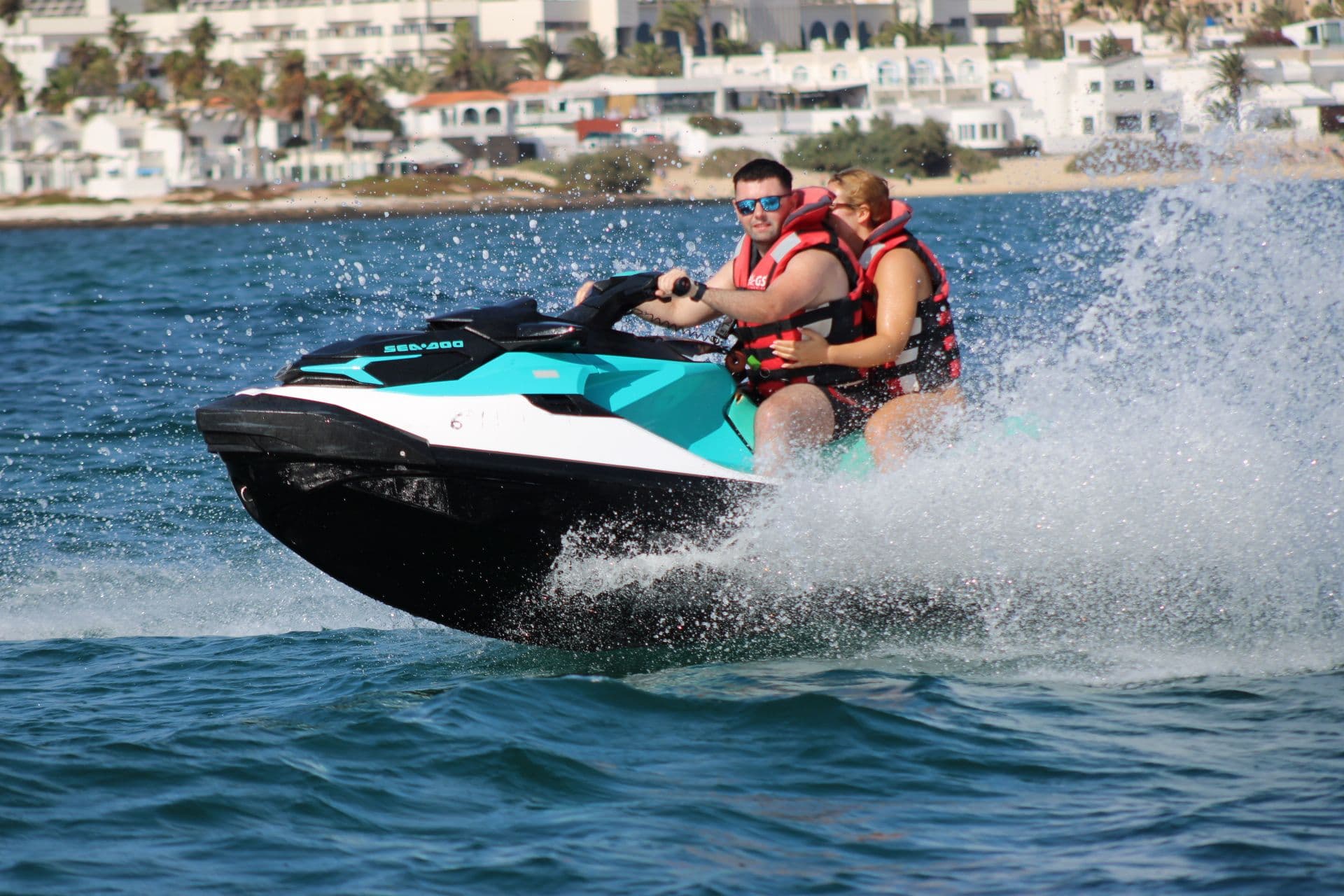 Couple on a turquoise jet ski throwing a wall of spray in front of the white houses of Corralejo