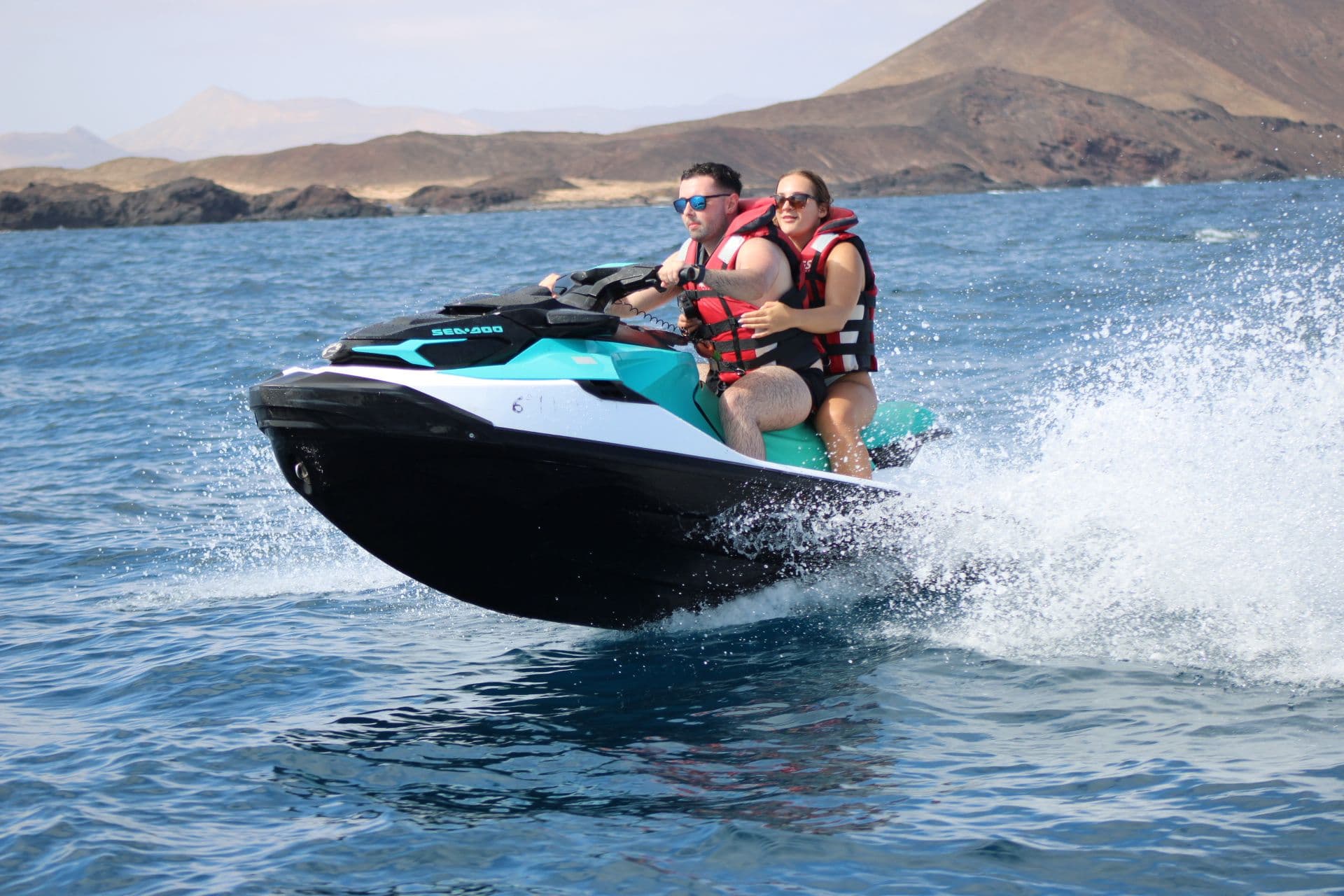 Couple riding a turquoise jet ski with the volcanic coast of north Fuerteventura behind