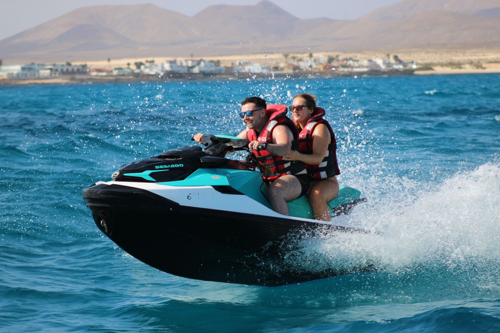Couple catching air off a wave on a turquoise Sea-Doo with the Corralejo coastline behind