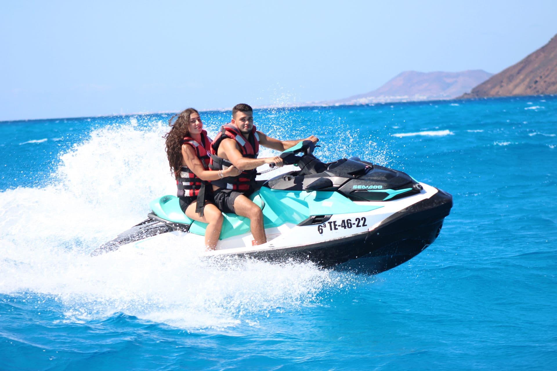 Couple jumping a wave on a turquoise and white Sea-Doo with the volcanic headland behind