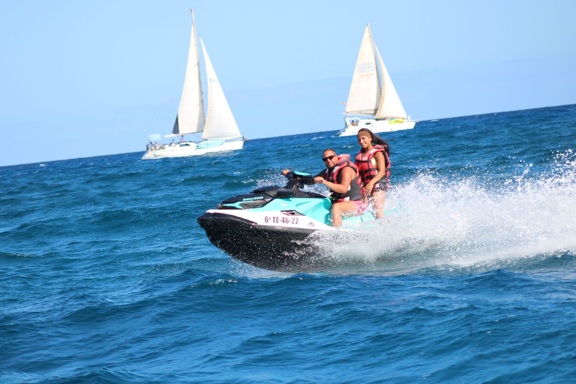 Couple riding a jet ski past two white sailboats in deep blue water off Corralejo
