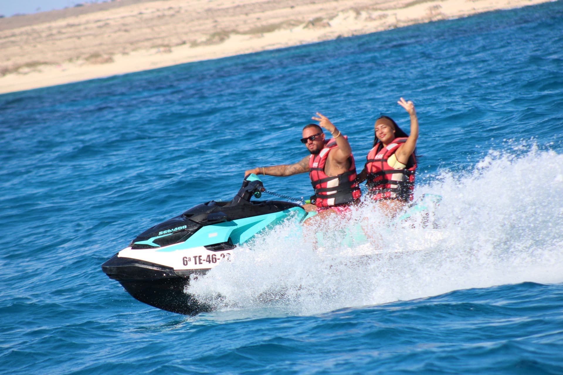 Couple waving from a turquoise jet ski as it carves through blue water near Corralejo