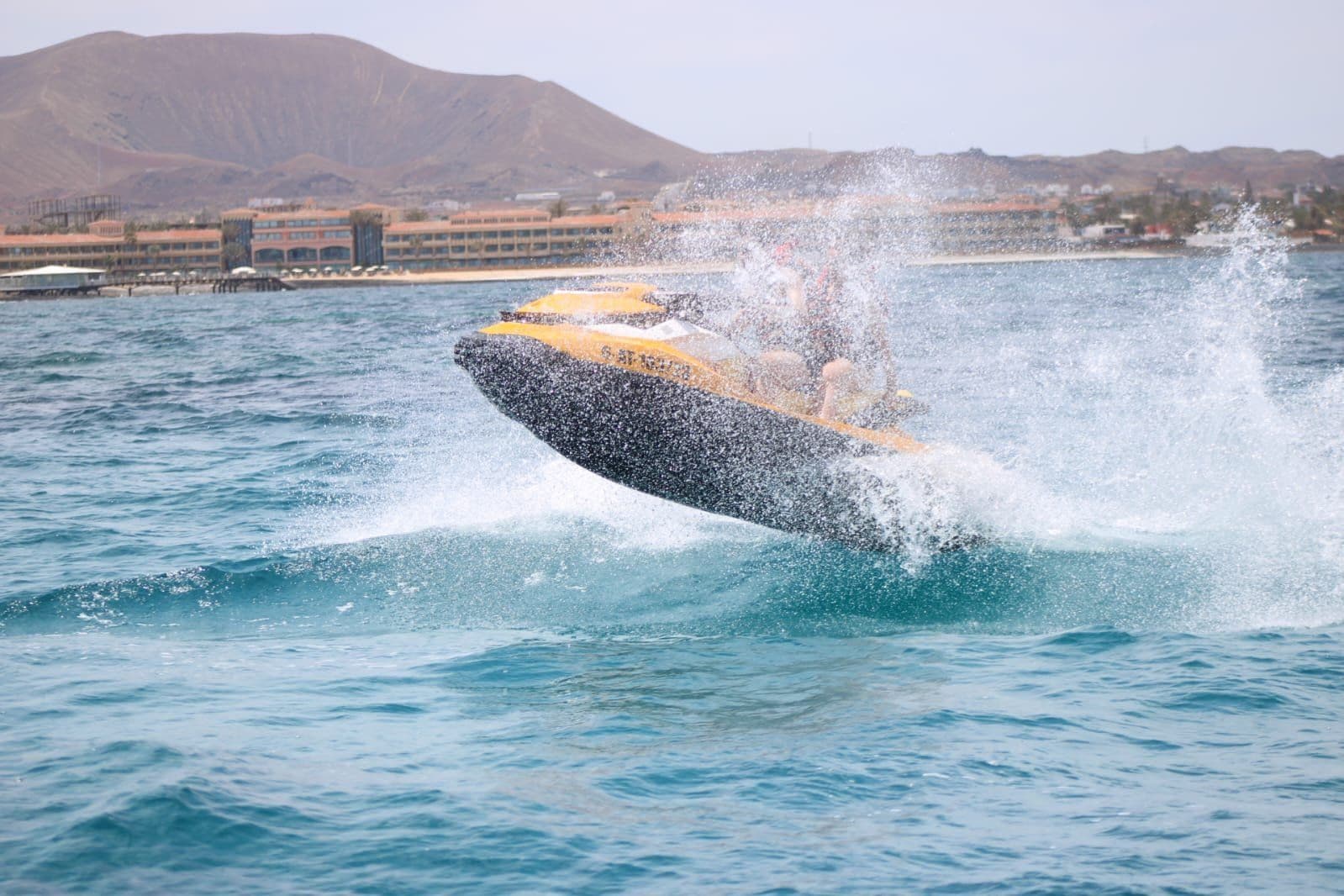 Couple in red life vests jumping a yellow jet ski near the white village houses of Corralejo, Fuerteventura