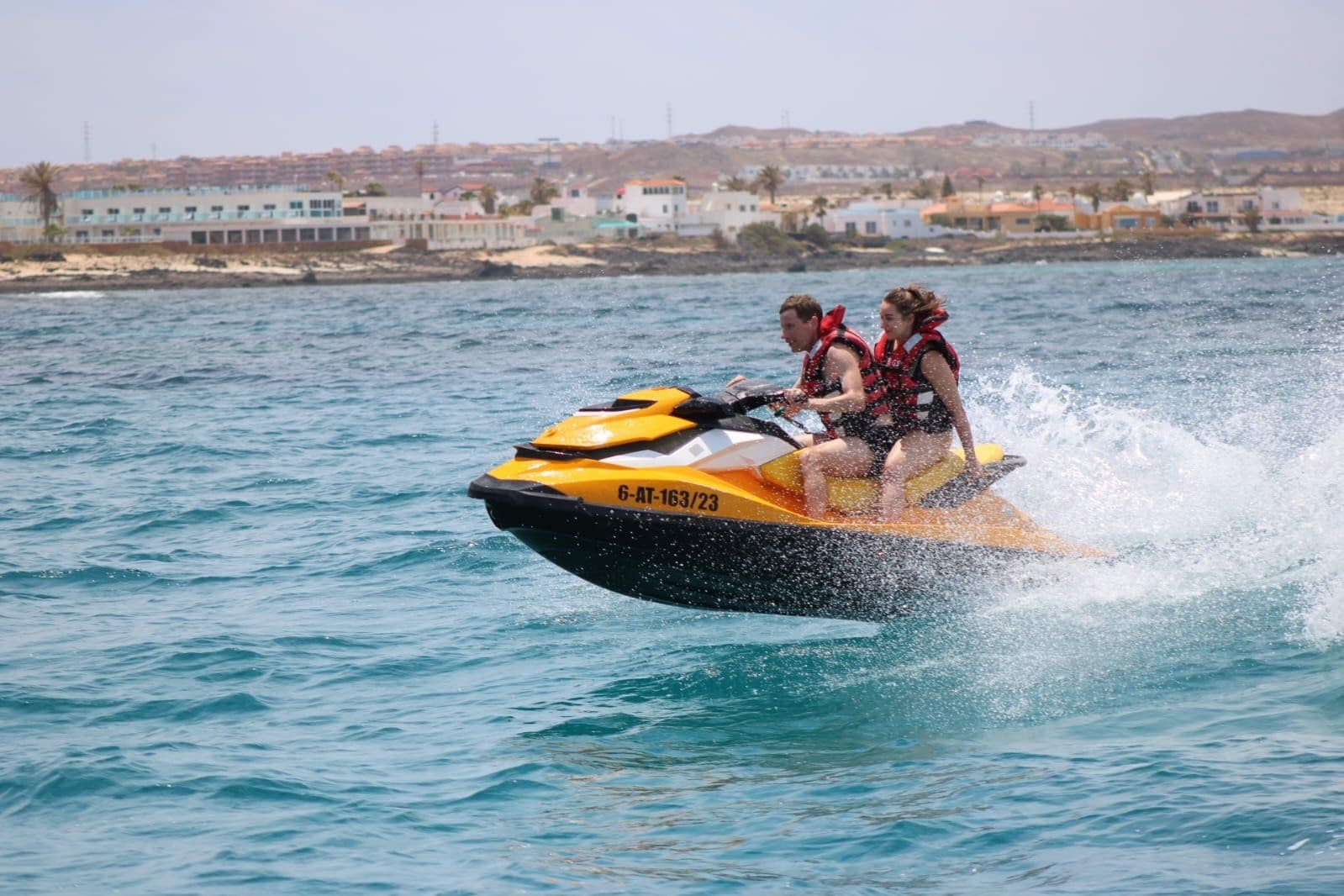 Smiling couple riding a yellow jet ski with the famous Corralejo sand dunes and Sahara-like beach in the distance