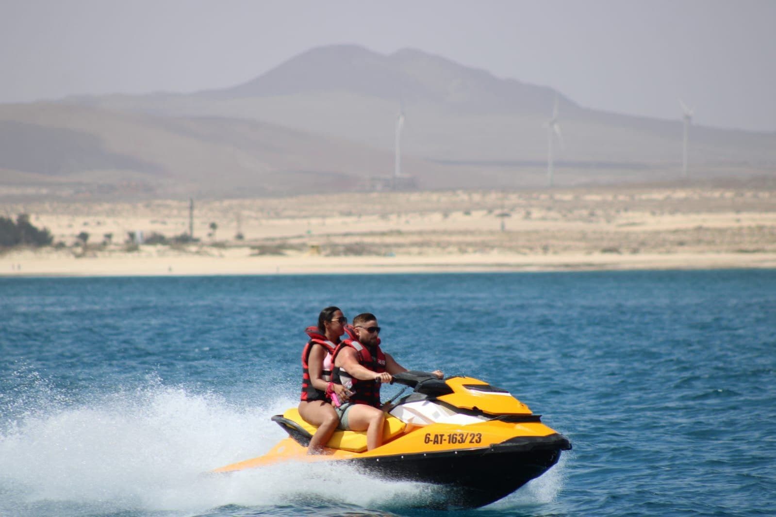 Couple on yellow jet ski with the desert mountains and iconic wind turbines of Fuerteventura in the distance