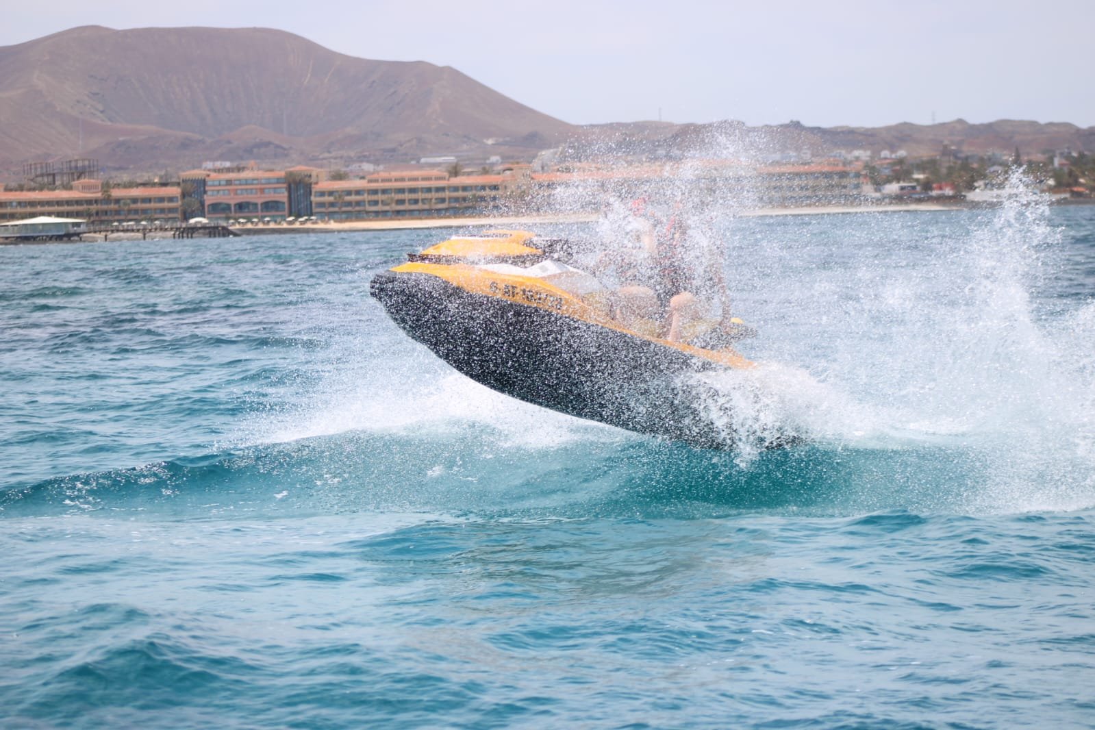 Couple in red life vests jumping a yellow jet ski near the white village houses of Corralejo, Fuerteventura