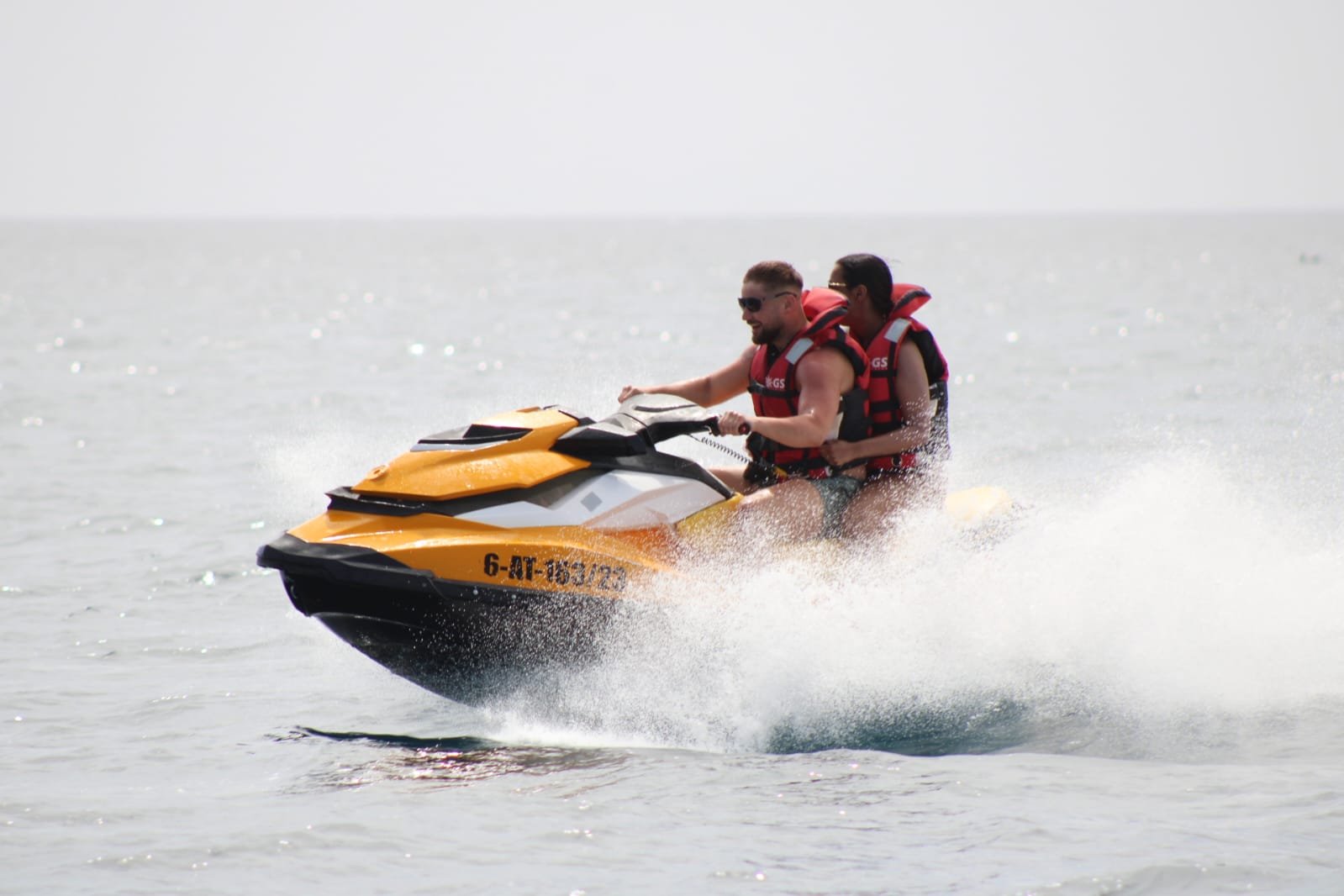 Couple on a black Sea-Doo jet ski jumping a wave with kitesurfers in the background near Corralejo