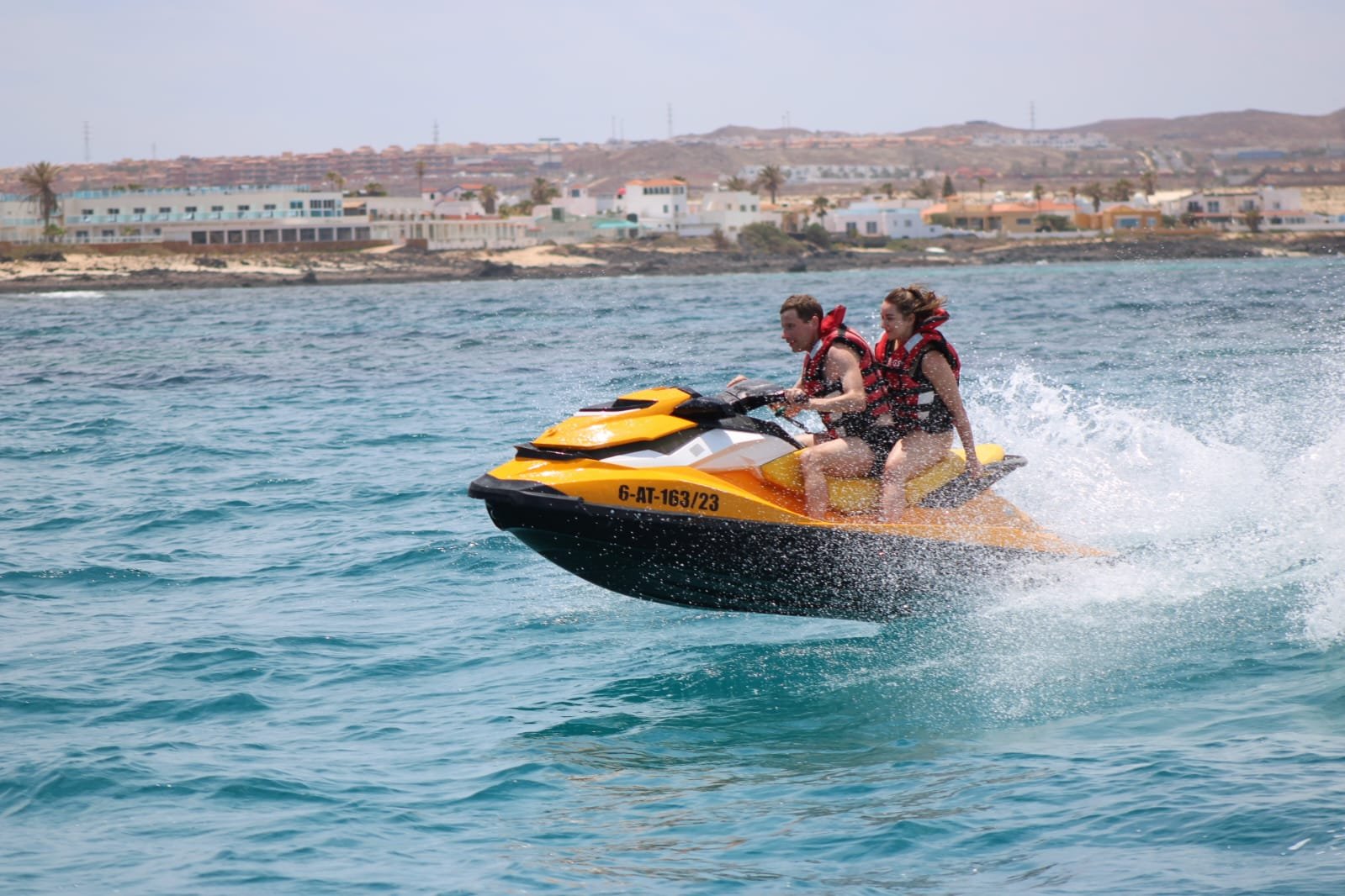 Smiling couple riding a yellow jet ski with the famous Corralejo sand dunes and Sahara-like beach in the distance