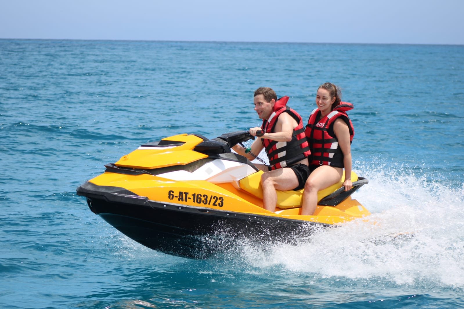 Couple on a yellow jet ski with the Punta Martiño lighthouse and Lanzarote mountains in the distance, off the coast of Fuerteventura