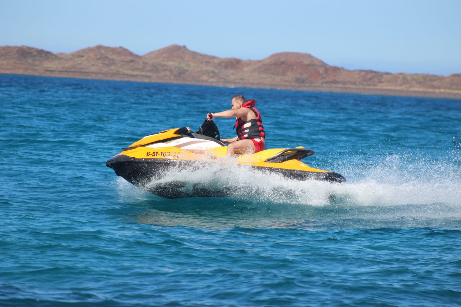 Couple in red life vests airborne on a yellow Sea-Doo jet ski near the rocky coastline of Fuerteventura