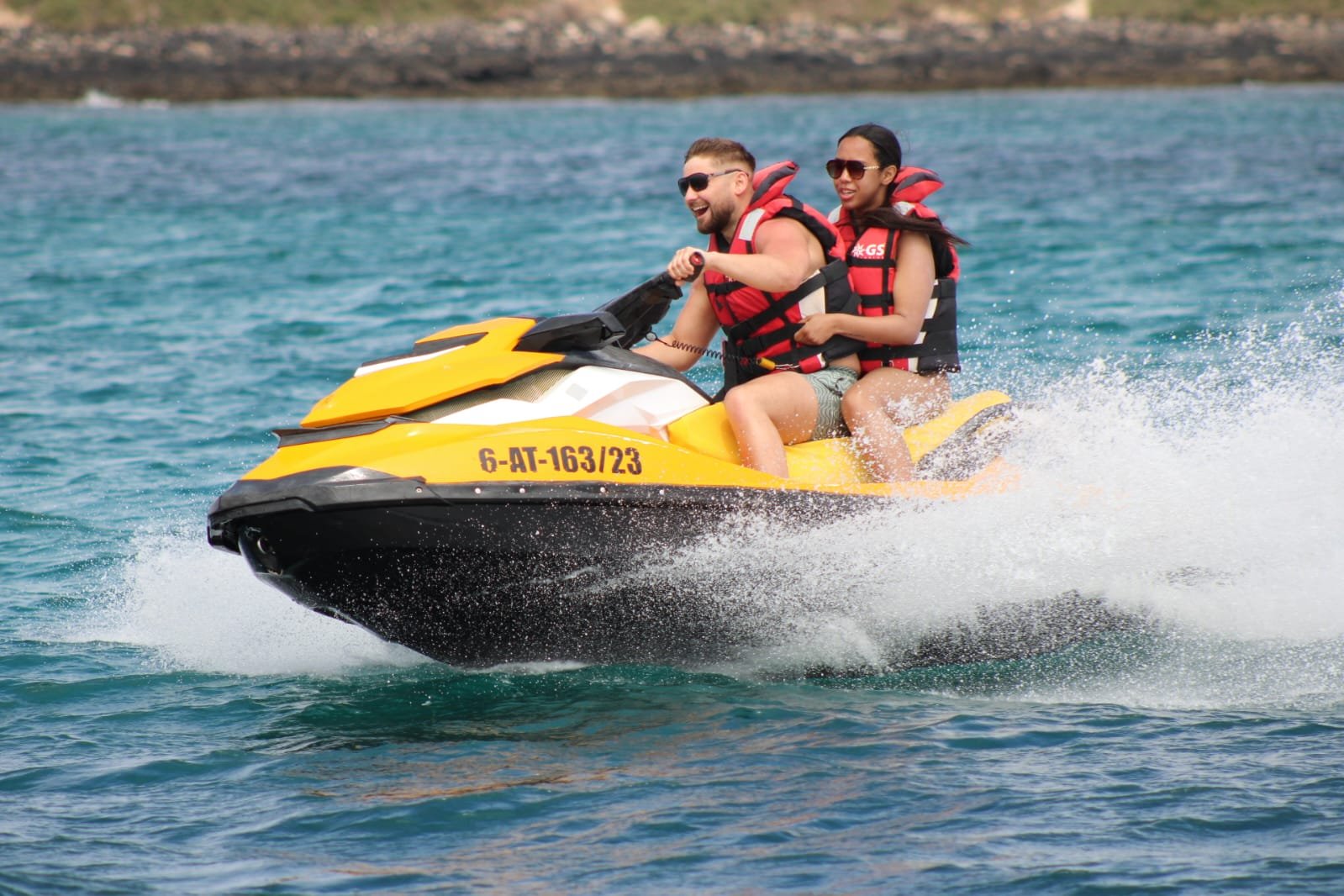 Couple wearing sunglasses airborne on a yellow Sea-Doo jet ski with the rocky coast of Fuerteventura behind