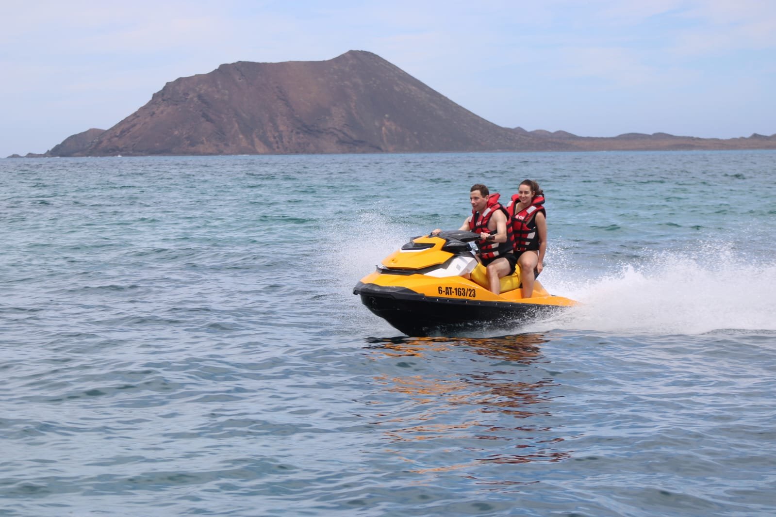 Smiling couple wearing red life vests riding a yellow Sea-Doo jet ski on the turquoise Atlantic waters near Corralejo, Fuerteventura
