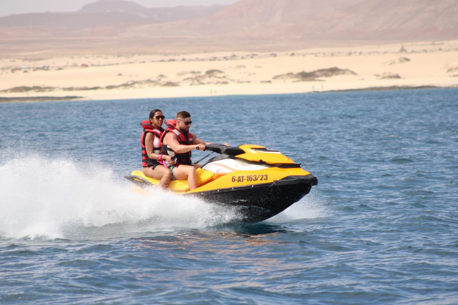 Couple riding a yellow jet ski past the bright white Corralejo sand dunes of Parque Natural
