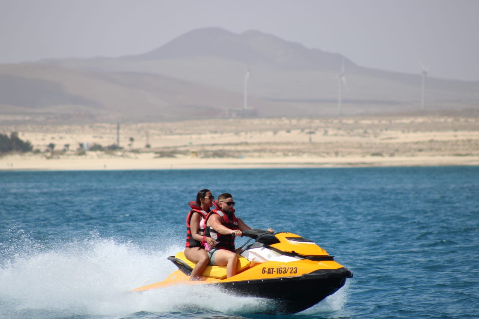 Couple on yellow jet ski with the desert mountains and iconic wind turbines of Fuerteventura in the distance