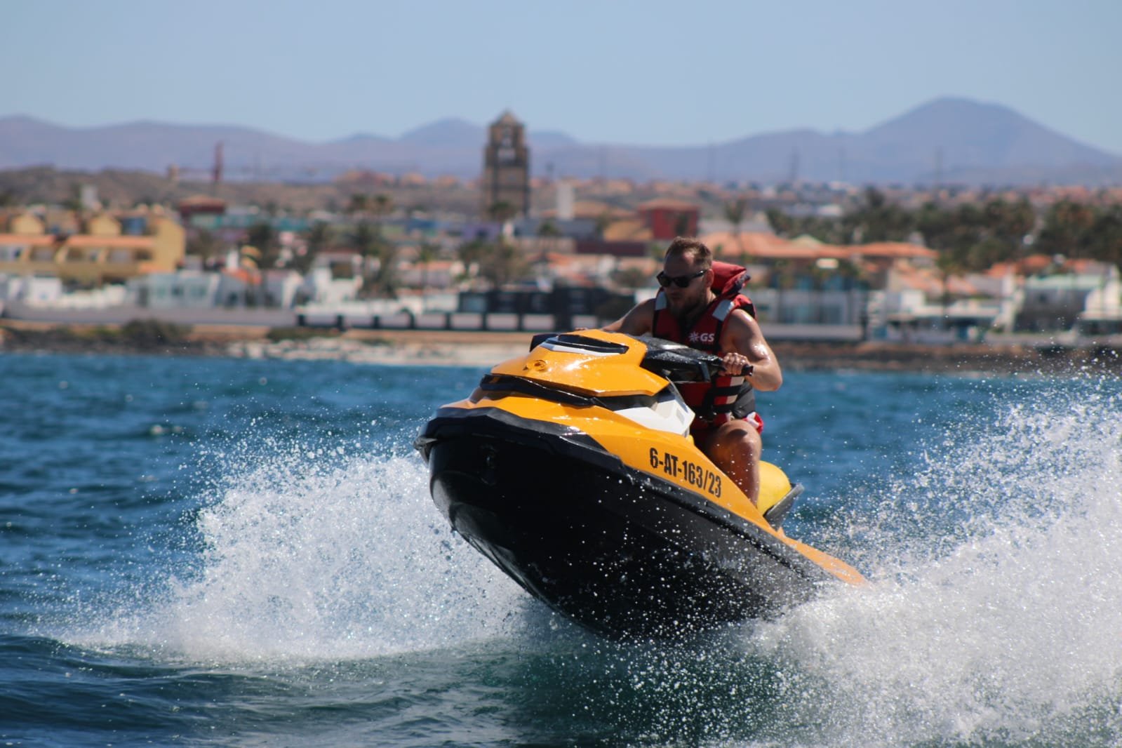 Solo rider jumping a yellow jet ski with the Corralejo beach hotels and mountains behind, mid-air