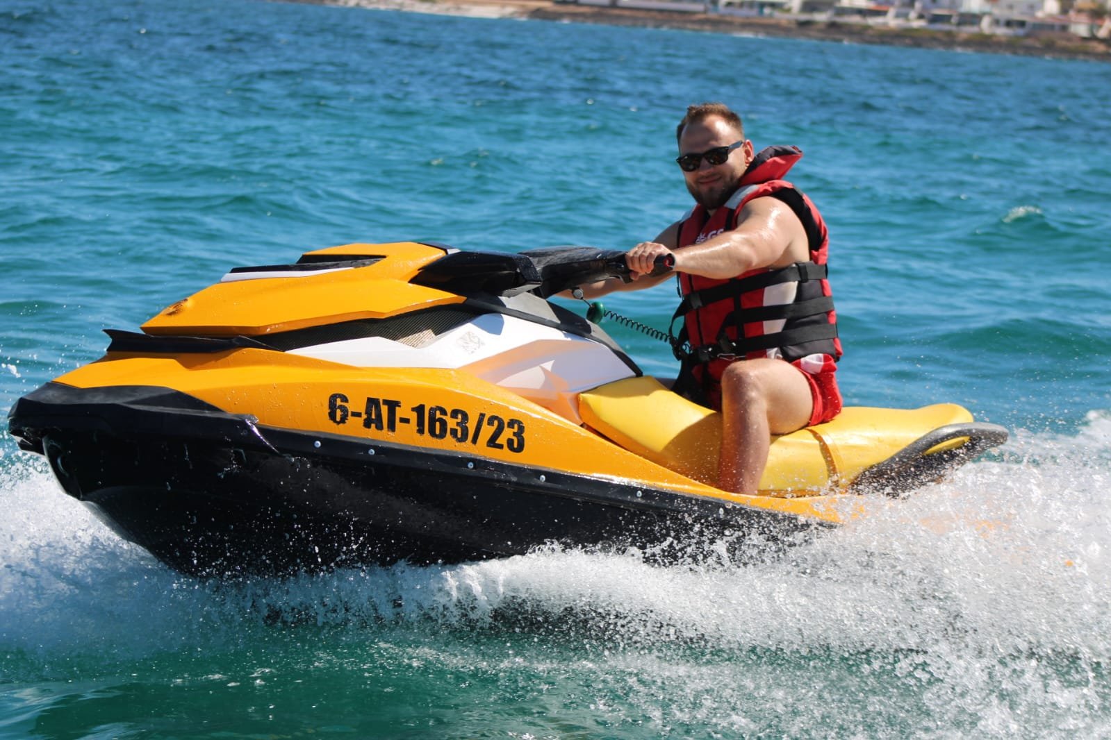 Solo rider on a yellow Sea-Doo jet ski with white Corralejo beachside houses and palm trees behind