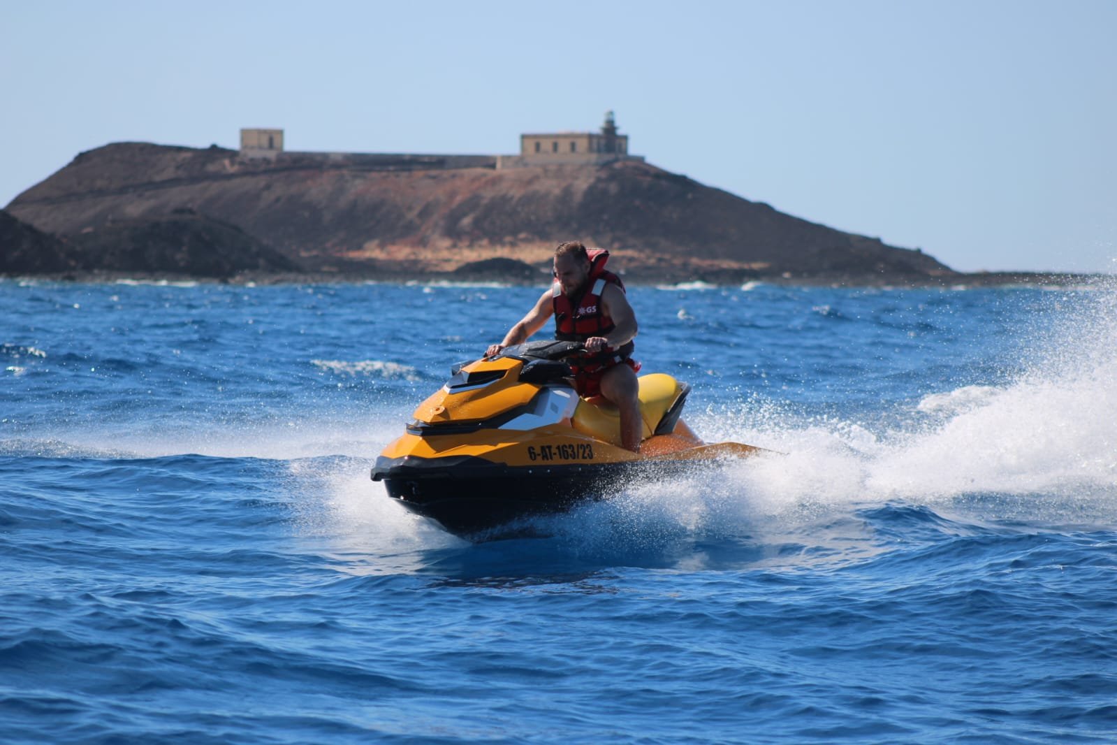 Solo rider in sunglasses on a yellow jet ski with the Corralejo town skyline and church tower in the distance
